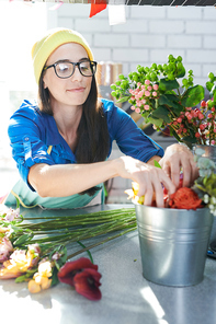 Portrait of modern young woman wearing glasses arranging flower compositions and smiling happily while enjoying work in sunlit shop, copy space