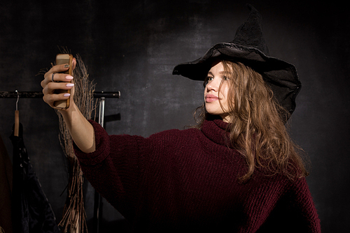 Smiling beautiful young lady with curly hair wearing witch costume standing against blackboard and taking selfie on smartphone in dark room