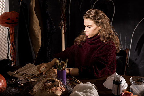 Serious concentrated beautiful young female mask designer with curly hair sitting at table and choosing paintbrush while preparing for work in dark design studio