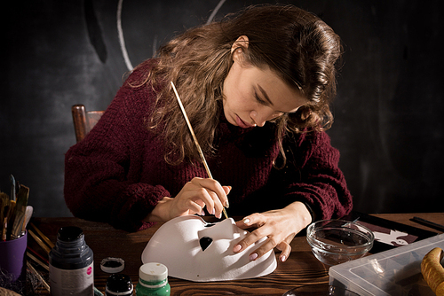 Serious concentrated attractive young woman in sweater sitting at wooden table and painting mask in dark workshop