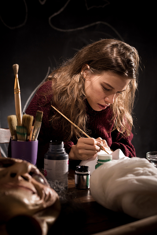 : Serious concentrated young female mask-maker with curly hair sitting at table and using paintbrush while working on design of Halloween mask in dark room