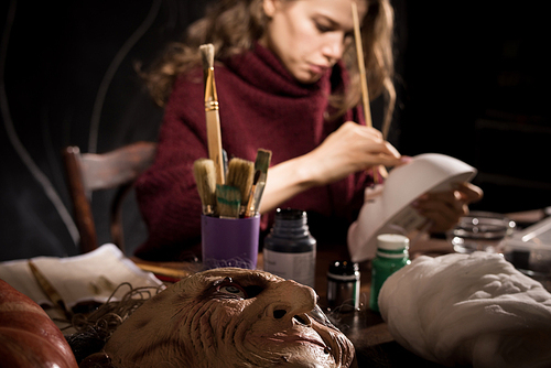 Close-up of busy young woman sitting at table and making realistic Halloween mask in workshop. Focus on witch face mask on table