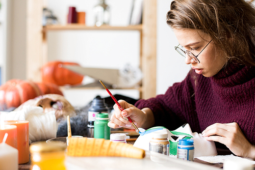Serious busy attractive young craftswoman in glasses sitting at table and elaborating design of Halloween mask, she painting mask in art studio