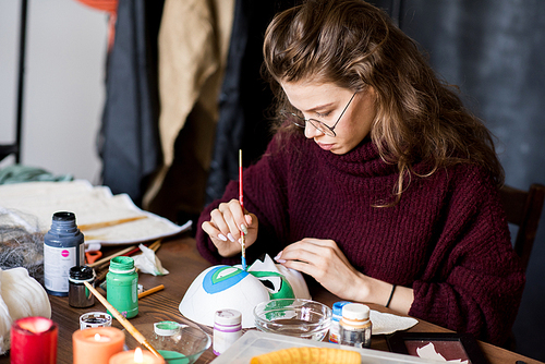 Serious skilled attractive young woman in glasses sitting at table and focusing on work while painting mask with paintbrush in own art studio