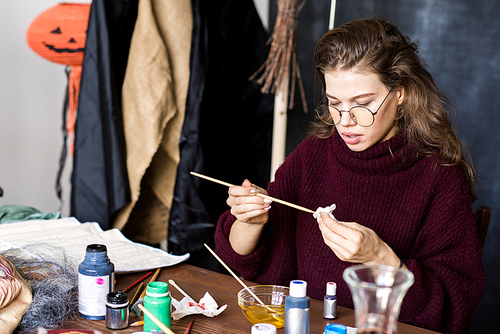 Serious concentrated young woman in round eyeglasses sitting at table and removing paint from brush using paper napkin while working in art studio at Halloween eve.