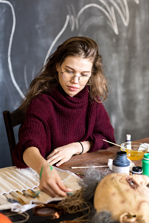 Serious pretty young female mask designer in glasses wearing warm sweater sitting at table and choosing paintbrush for work in art studio