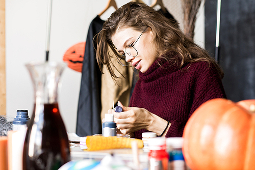 Positive attractive young woman in glasses sitting at table and reading label on paint jar while preparing for mixing colors in art studio