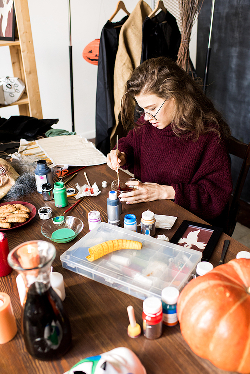 Serious busy girl in glasses sitting at table and mixing paints in bowl while preparing for work in art studio, she making Halloween costume