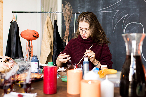 Serious concentrated young woman in glasses preparing for painting in workshop: she adding red paint in bowl using syringe and mixing it with paintbrush