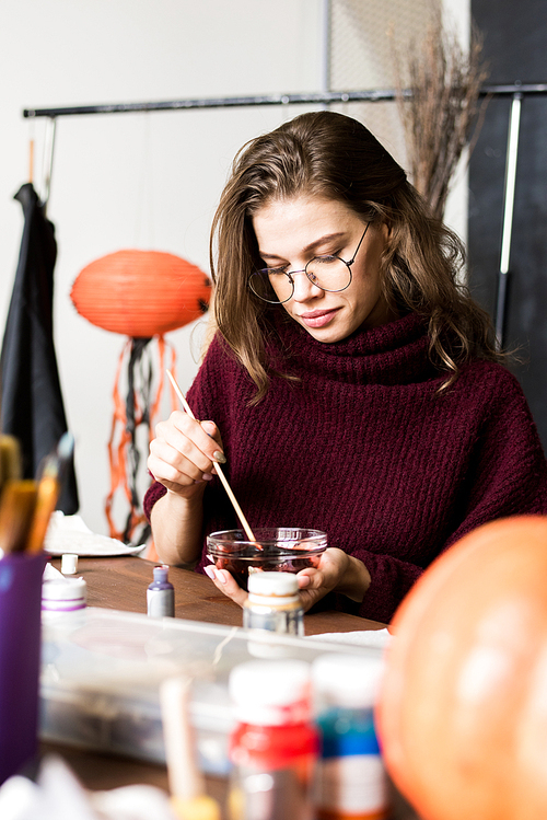 Concentrated young creative artist in warm sweater sitting at table and mixing paint in bowl while preparing for work on Halloween decoration in art studio