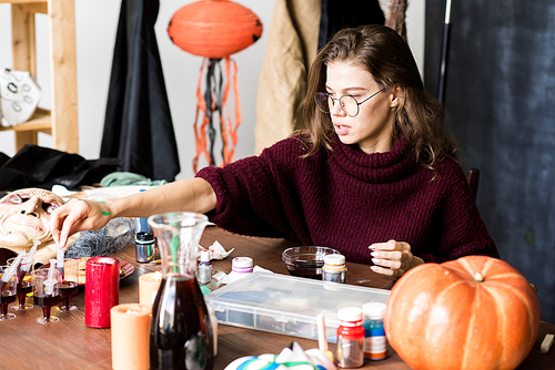 Serious confident modern young woman in glasses sitting at table with Halloween decorations and art tools and working on Halloween design using paint of blood color in workshop
