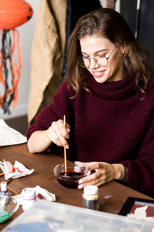 Happy excited creative young female designer in glasses sitting at table and mixing paint to make color of blood for Halloween decoration, she enjoying work in art studio