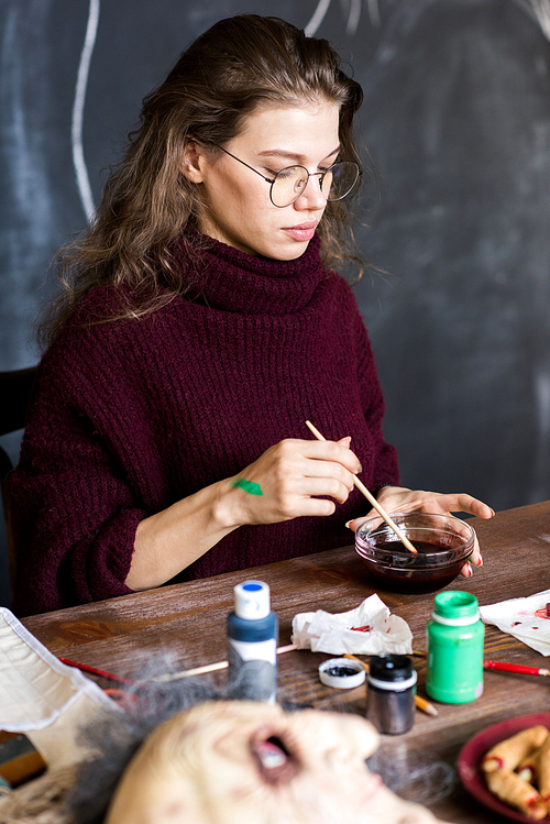 Serious concentrated young woman in glasses wearing warm knitted sweater sitting at table with art tools and mixing paints with brush in bowl while working on Halloween design in art studio.