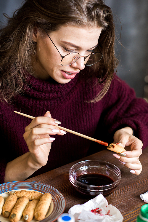 Positive attractive young creative lady in glasses sitting at table and painting Halloween cookies with food coloring in workshop