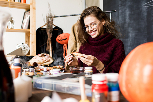 Excited attractive young lady in glasses sitting at table full of art equipment and making Halloween cookie more realistic while having fun in own studio