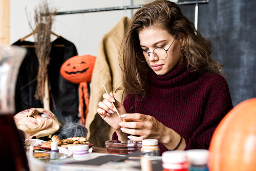 Serious confident creative young female designer in glasses sitting at table and paining food for Halloween in workshop, making witch finger cookie