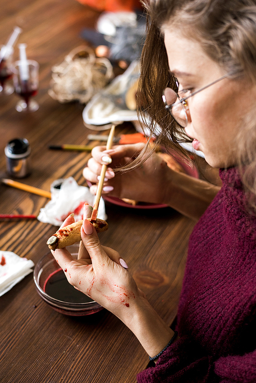 Close-up of concentrated female food designer immersed into work sitting at dirty wooden table and painting witch finger cookie with food dye of blood color in workshop.