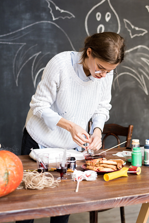Serious concentrated young woman in white sweater standing at table and drawing up liquid paint in syringe while preparing for decorating gauze in workshop, Halloween decor