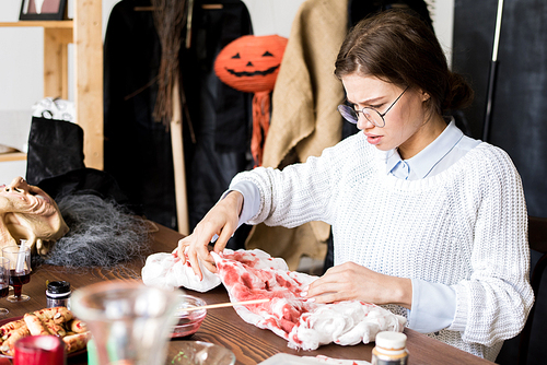 Serious concentrated young woman in glasses sitting at table and making bloody stains on Halloween costume, she checking paint on gauze in workshop