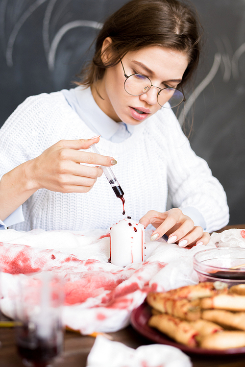Serious concentrated young woman in glasses sitting at table and painting candle for Halloween, she dropping red paint from syringe in workshop