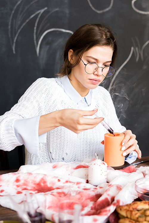 Serious concentrated beautiful girl in round glasses sitting at table with clothes in red paint and adding paint into candle while working on Halloween decoration in workshop.