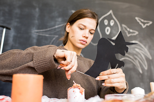 Serious busy girl in sweater sitting at table against blackboard with drawn ghost and cutting witch out of black paper while working on Halloween decoration in workshop.