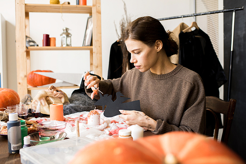 Serious concentrated young craftswoman in casual sweater sitting at table and cutting out paper witch using scissors, she making Halloween decoration in creative workshop