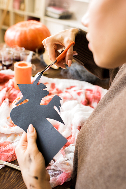 Close-up of busy woman sitting at table and carefully cutting out paper witch using scissors above table with bloody gauze in workshop