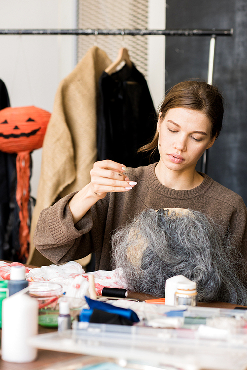 Serious concentrated young woman in casual sweater sitting at table with craft tools and decoration stuff and using sewing needle while attaching hair to Halloween mask in workshop.