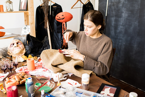 serious busy young woman in brown sweater sitting at desk full of halloween stuff and tools and adding colorful  leaves while sewing burlap in workshop.