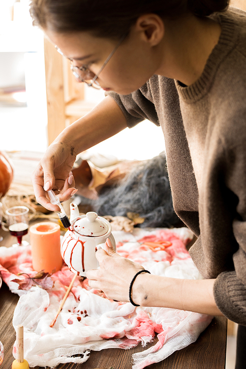 Serious busy young woman in casual sweater standing at desk and using syringe while painting teapot with artificial blood above dirty fabric in workshop, making Halloween decor