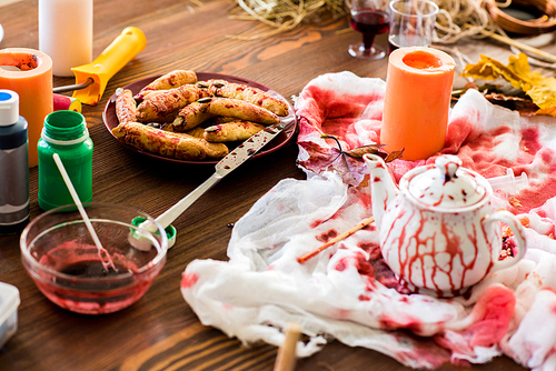 Creative space for decorating: close-up of white fabric with bloody stains, teapot, candle, gouache jars and plate of witch finger cookies on wooden table