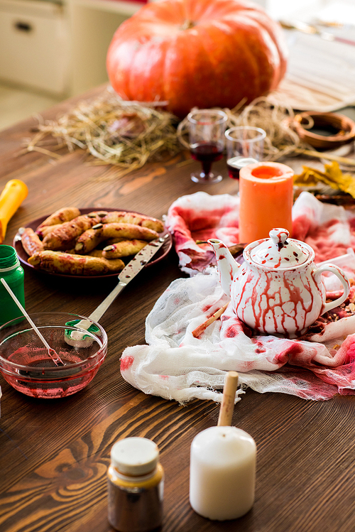Workplace of Halloween designer: close-u of dirty wooden table with bloody fabric with teapot, bowl of liquid paint, bloody knife leaning on plate with witch finger cookies in workshop