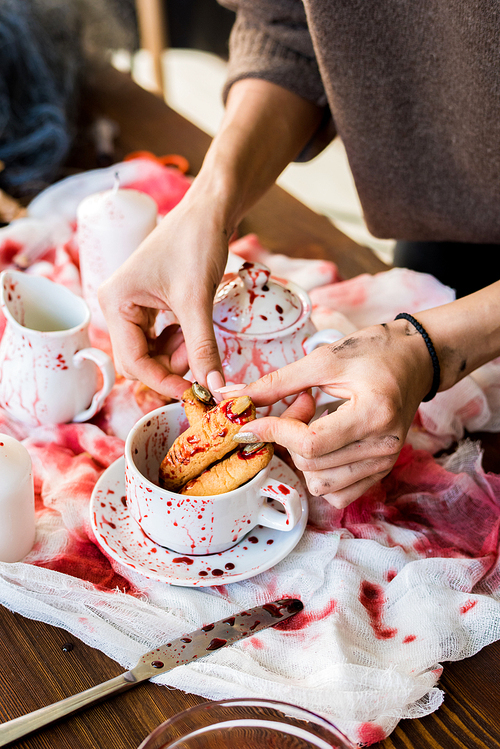 Close-up of unrecognizable girl standing at table with tea crockery and gauze and adjusting witch finger cookie in cup while making Halloween decor  in workshop.