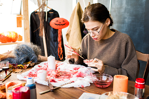 Serious concentrated attractive young craftswoman in glasses sitting at table with Halloween decoration and art equipment and adding paint on toy jaw in workshop