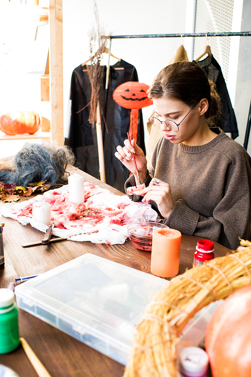 Serious modern young female designer in eyeglasses sitting at table and painting jaw model with red paint while making Halloween costume in workshop