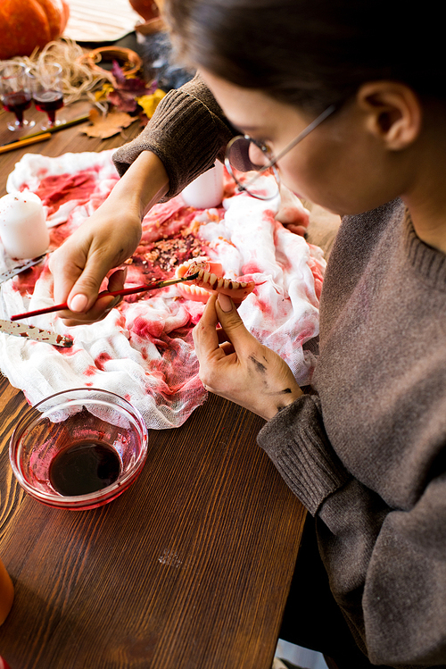 Close-up of concentrated young woman sitting at wooden table with bloody gauze and painting jaw model with small paintbrush while creating spooky costume for Halloween in workshop.
