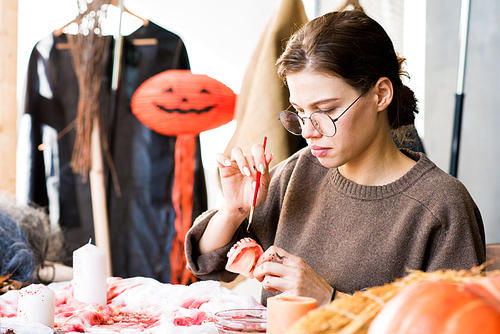 Serious beautiful young lady in glasses sitting at table full of Halloween stuff and painting toy jaw with paint of blood color while working on costume in workshop.