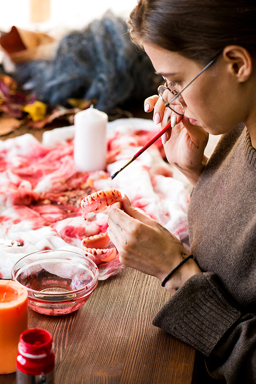 Close-up of serious concentrated young woman in glasses sitting at desk and applying paint of blood color while making dracula teeth in workshop