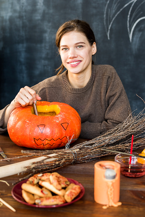 Happy attractive girl in brown sweater sitting at wooden table with Halloween decoration like pumpkin, broom and witch finger cookies and  cheerfully in workshop