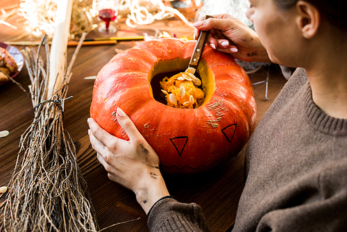 Close-up of concentrated woman in sweater sitting at table with broom and scooping out pumpkin seeds while making jack-o-lantern in workshop