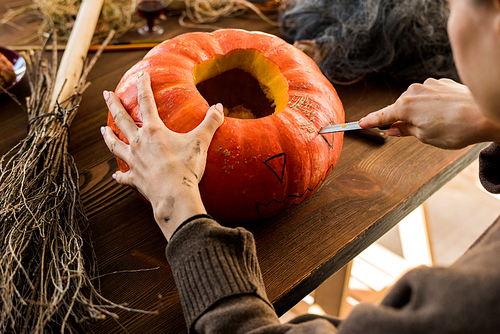 Close-up of unrecognizable woman sitting at table and cutting eye on pumpkin while making jack-o-lantern in workshop