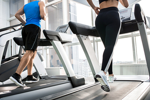 Mid-section back view of two fit young people, man and woman, running on treadmills facing windows in modern gym, copy space