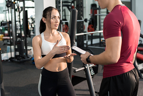 Portrait of young woman talking to personal fitness coach in modern gym, copy space
