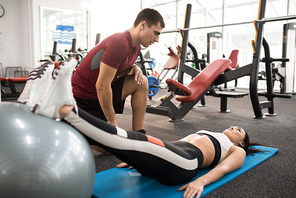 Full length  portrait of muscular fitness coach helping young woman doing exercises on mat in modern gym