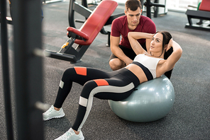 High angle portrait of muscular coach helping young woman doing exercises on fitness ball  in modern gym, copy space
