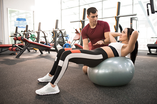 Side view portrait of muscular coach helping young woman doing exercises on fitness ball  in modern gym, copy space