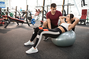 Portrait of muscular coach helping young woman doing exercises on fitness ball  in modern gym, copy space