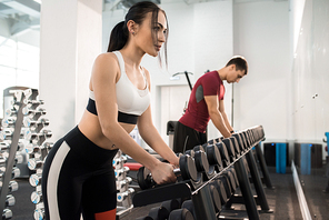 Side view portrait of beautiful young woman picking up dumbbells from equipment rack in modern gym, copy space