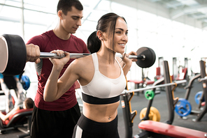 Portrait of fitness coach helping beautiful young woman training with barbell in modern gym, copy space